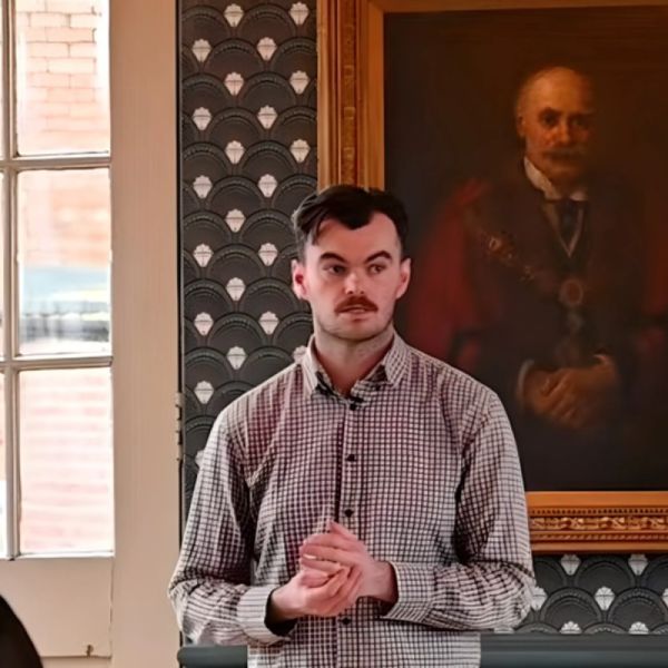 David Stubbs, standing with his hands clasped, in a room with patterned wallpaper and a framed portrait behind him.